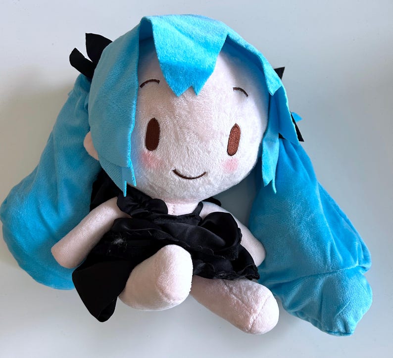 May include: A plush doll with a white face, pink cheeks, and brown eyes. The doll has long, light blue hair and is wearing a black dress. The doll's arms and legs are a light pink color. The doll is a cartoon character.