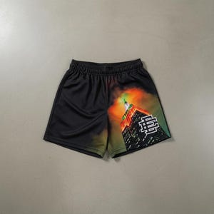 Puede incluir: Pantalones cortos de malla negros con un gráfico colorido del Empire State Building. Los pantalones cortos tienen una cintura elástica y un logotipo impreso en la pierna derecha. El gráfico incluye tonos de naranja, verde y rojo.