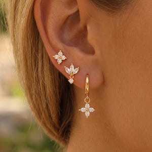 Op de afbeelding: Een set van drie goudkleurige oorbellen met heldere, marquise-geslepen stenen in bloemmotieven. De bovenste oorbel is een knopje met vier blaadjes, de middelste heeft drie blaadjes en de onderste is een kleine ring met een vierbladig hangertje.