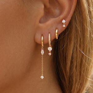 May include: A set of four gold-toned earrings worn in one ear. The collection includes a small plain hoop, a hoop with a dangling chain and marquise-cut stone, a hoop with a pavé-set band and round stone, and a small stud with two tiny stones.