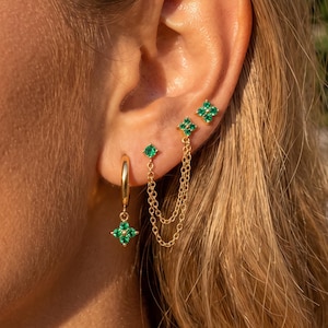 May include: A gold hoop earring with a dangling four-pointed green stone charm, paired with a double-chain earring featuring two four-pointed green stone studs connected by two delicate gold chains, all worn in a multi-pierced ear arrangement.