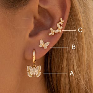 Può includere: Un set di tre orecchini a farfalla color oro con pietre pavé bianche. L'articolo A è un orecchino a cerchio con ciondolo a farfalla, l'articolo B è un orecchino a lobo a farfalla e l'articolo C è un orecchino a rampicante a farfalla.