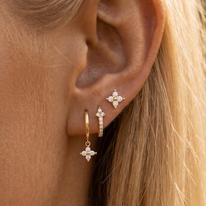 May include: A set of three gold-toned earrings featuring clear stones. One earring is a small hoop with a dangling four-petaled flower charm, one is a small hoop with a row of stones, and one is a stud earring shaped like a four-petaled flower.