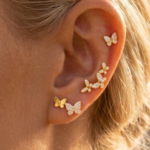 Può includere: Un set di orecchini a farfalla color oro con piccole pietre trasparenti. La collezione include un orecchino a lobo, un orecchino a rampicante con più farfalle e un paio composto da una farfalla dorata e una decorata con pietre incastonate.