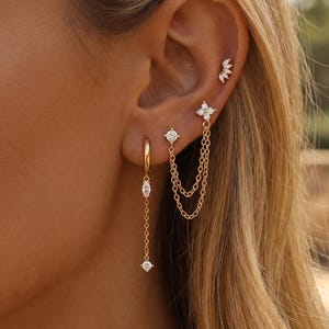 May include: A set of gold-toned earrings featuring a hoop with a dangling chain and marquise-cut stone, a stud with a double-chain drape connecting to a flower-shaped stud, and a small curved stud with multiple tiny stones in the upper ear cartilage.