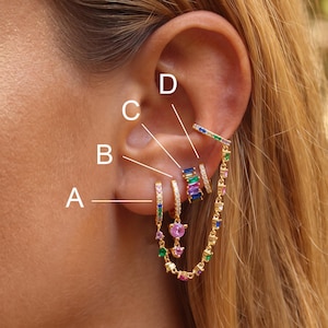 May include: A set of gold-tone earrings featuring four distinct pieces labeled A, B, C, and D. The jewelry includes small hoops with multi-colored baguette and round-cut stones, a pink circular stone charm, and a long chain connecting the pieces.