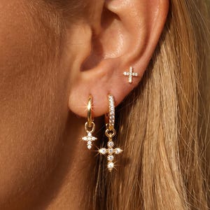 May include: A set of gold-toned earrings featuring three distinct pieces: a small cross stud earring, a small hoop earring with a dangling cross charm, and a larger hoop earring with a pavé-set cross charm, all adorned with clear crystals.