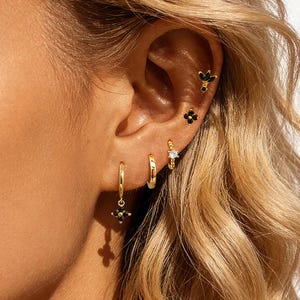 May include: An ear displaying various gold earrings, including small hoops and studs. Some earrings feature black flower-shaped designs and a clear stone. The ear is framed by wavy blonde hair, creating a contrast with the gold jewellery.