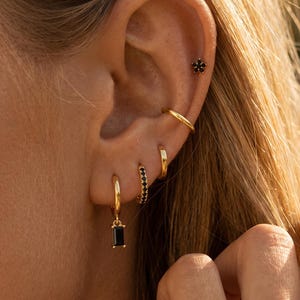 May include: A set of gold-toned earrings featuring a star-shaped stud with black stones, two plain gold hoops, a hoop with black stones, and a hoop earring with a dangling rectangular black stone charm. The jewelry is worn in multiple ear piercings.