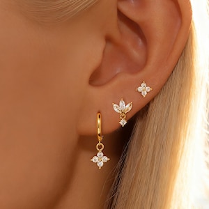 以下が含まれることがあります： 3つの異なるゴールドのイヤリングで飾られた人の耳のクローズアップ。イヤリングは、花柄と葉の模様の透明で輝く宝石が特徴です。1つのイヤリングは小さなスタッド、もう1つはぶら下がっているデザイン、3つ目は花のチャームが付いた小さなフープです。