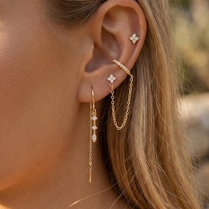 Può includere: Un set di orecchini e ear cuff in oro composto da un piccolo perno a forma di fiore a quattro petali con pietre chiare, una catenina sottile che collega il cuff a un perno a fiore e un lungo orecchino pendente con due piccoli fiori e una pietra.
