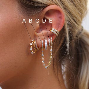 Op de afbeelding: Een close-up van een oor versierd met meerdere gouden oorbellen. De oorbellen omvatten creolen, studs en een kettingontwerp, allemaal versierd met heldere, sprankelende edelstenen. De sieraden zijn geplaatst tegen de achtergrond van de huid en het haar van de persoon.