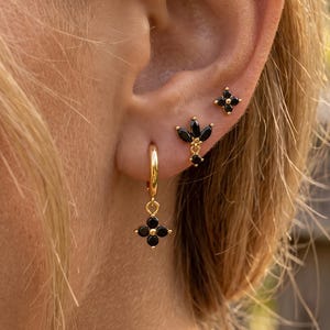以下が含まれることがあります： 黒い石があしらわれたゴールドトーンのピアス3点セット。一番下のピアスは4枚花びらのチャームが付いた小さなフープ型。真ん中のピアスはマーキスカットのクラスター型で、一番上のピアスは小さな四芒星のスタッド型です。