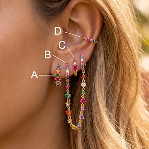 May include: A gold-toned ear stack featuring four distinct pieces labeled A, B, C, and D. Piece A is a small multi-colored hoop. Piece B is a hoop connected by a chain to a larger hoop. Piece C is a hoop with a rectangular pink stone. Piece D is a thin multi-colored ear cuff.