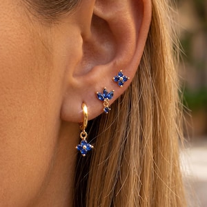 Könnte beinhalten: Ein Set aus drei goldfarbenen Ohrringen mit dunkelblauen Steinen. Der obere Ohrring ist ein Stecker in Form einer vierblättrigen Blume, der mittlere ein dreiblättriger Stecker und der untere ein kleiner Goldring mit einem passenden Blumenanhänger.