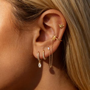 May include: An ear is adorned with gold-coloured earrings. The set includes a bee stud, a bee ear cuff with a chain, and two hoop earrings, one with a dangling teardrop-shaped gem. The hoops are encrusted with small, clear stones.