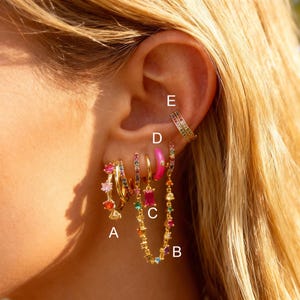 Könnte beinhalten: Eine Sammlung farbenfroher Ohrringe, darunter goldene Creolen mit Regenbogen-Edelsteinen, ein pinkfarbener Emaille-Reifen und eine Kette aus bunten Edelsteinen. Die Ohrringe werden an einem Ohr präsentiert und zeigen verschiedene Stile.