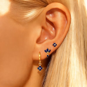May include: A close-up of an ear adorned with three sets of gold-colored earrings. The earrings feature blue gemstones in floral and leaf-like designs. One earring is a small stud, another is a dangle, and the third is a small cluster.