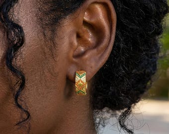 May include: A gold hoop earring featuring a geometric pattern of inlaid orange and green stones. The earring has a thick, rounded band design with a repeating diamond and chevron motif, showcasing a polished metallic finish and vibrant gemstone accents.