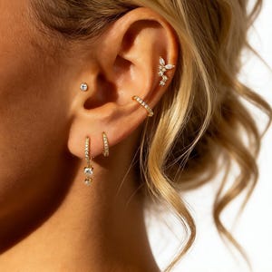 May include: Close-up of a person's ear adorned with multiple gold earrings. The ear features a small stud, a hoop earring, a dangling earring with a gem, and a cuff earring. A floral-shaped earring is also visible. The earrings are embellished with small, sparkling stones.