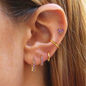 May include: An ear featuring several gold earrings. A butterfly stud with purple gemstones is at the top, alongside a gold hoop and two small hoops with purple gems. A gold ear cuff is also visible.