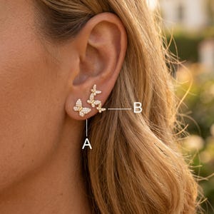 May include: Gold-toned butterfly earrings featuring small clear stones. One earring is a single butterfly labeled A, and the other is a cluster of three butterflies labeled B. The jewelry is worn on the earlobe, showcasing a delicate design.