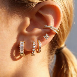 May include: A collection of gold-toned earrings and ear cuff. The earrings include a pair of baguette-cut hoop earrings, a pair of small hoop earrings with three clear stones, a pair of small hoop earrings with clear baguette stones, and a flower-shaped stud earring.
