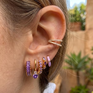 May include: A gold-toned ear cuff with clear stones sits on the upper ear cartilage. Below, four gold-toned hoop earrings are worn: one with purple baguette stones, one with a purple round stone, one with purple round stones, and one with a purple enamel finish.