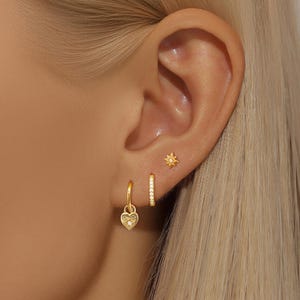 Puede incluir: Primer plano de una oreja adornada con tres pendientes dorados. Los pendientes incluyen un dije en forma de corazón, un aro pequeño con piedras transparentes y un pendiente en forma de estrella. Los pendientes están sobre la piel y el pelo rubio.
