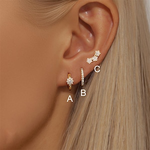 Può includere: Tre orecchini d'oro sono mostrati su un orecchio. Il primo è un piccolo cerchio con un design a forma di fiore. Il secondo è un piccolo cerchio con una fila di piccole pietre. Il terzo è un orecchino curvo con tre disegni a forma di stella.