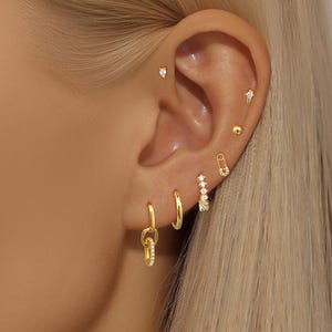 May include: A close-up of a person's ear adorned with multiple gold earrings. The earrings include small studs, hoops, and a safety pin design, all embellished with sparkling gemstones. The jewelry is set against the backdrop of the ear and blonde hair.