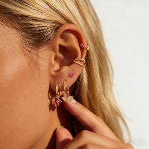 May include: A collection of gold-toned earrings and ear cuffs with pink gemstones. The earrings include small studs, hoop earrings with a pink flower design, and a fan-shaped charm. The ear cuffs and hoops have a textured design.