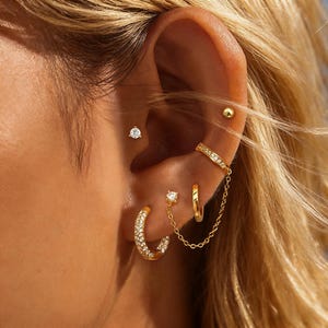 May include: A close-up of an ear adorned with multiple gold earrings. The earrings include a small stud with a clear stone, a hoop earring with clear stones, a chain connecting two earrings, and a gold ear cuff.