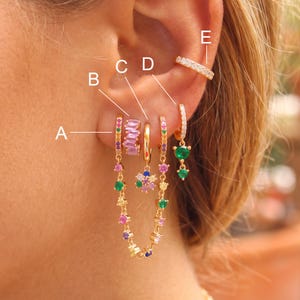 Puede incluir: Una colección de pendientes dorados etiquetados de la A a la E. A es un aro de piedras multicolor con cadena colgante, B es un aro de baguette rosa, C es un aro dorado liso, D es un aro con dije de flor y E es un pendiente de presión.