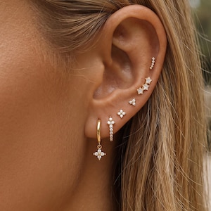 Op de afbeelding: Een collectie goudkleurige oorbellen met heldere stenen, waaronder een oorring met een vierpuntige sterhanger, een kleine ring met een verticale stenen balk en diverse oorknopjes in de vorm van sterren en clusters van kleine steentjes.