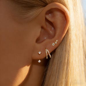 Könnte beinhalten: Nahaufnahme eines Ohrs, das mit mehreren goldenen Ohrringen geschmückt ist. Die Ohrringe umfassen kleine Ohrstecker, einen gebogenen Reifen und eine Ansammlung von drei Steinen. Der Schmuck funkelt auf der Haut und erzeugt einen zarten und eleganten Look.