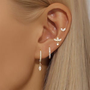 May include: A close-up of an ear adorned with multiple gold earrings. The earrings include small hoops, a dangling teardrop-shaped charm, and a floral design with clear gemstones. The ear is set against a backdrop of blonde hair.