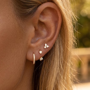 May include: A gold hoop earring with small clear stones and three additional gold stud earrings featuring clear stones. One stud is a three-stone cluster, while the other two are single stones, all worn in a series of piercings on the earlobe.