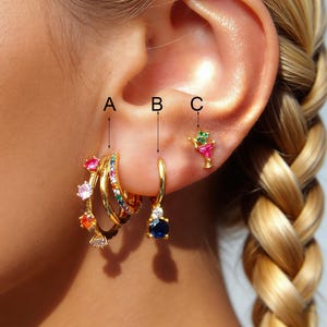 May include: Gold earrings with colorful gemstones. The earrings include a set of three hoops with rainbow gems, a single hoop with a blue gem, and a cocktail-shaped stud with pink and green gems. The earrings are displayed on an ear.