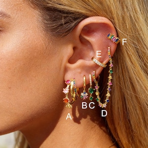 Può includere: Orecchini dorati con pietre preziose colorate. Gli orecchini includono modelli a cerchio, design pendenti e orecchini a polsino. Le pietre preziose sono di varie forme e colori, tra cui rosa, verde, blu e giallo. I gioielli sono esposti su un orecchio.