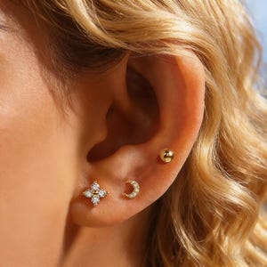 May include: A close-up of an ear adorned with three gold earrings. One is a small ball stud, another is a crescent moon shape with small clear stones, and the third is a four-petal flower shape with clear stones.