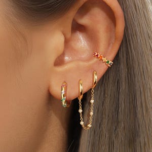 Puede incluir: Juego de pendientes y puño de oreja dorados. El conjunto incluye un par de pendientes de aro pequeños con gemas coloridas, un solo pendiente de aro y un pendiente de cadena. El puño de oreja presenta una fila de gemas coloridas en forma de estrella.