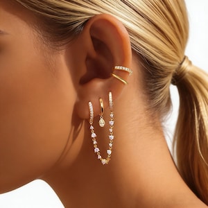 May include: A collection of gold-toned earrings and ear cuffs. The earrings include a pair of hoop earrings, a small dangling earring, and a chain of small, round, white gemstones. The ear cuffs are adorned with small, clear gemstones.