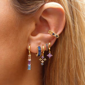 May include: A collection of gold-toned earrings and ear cuff. The earrings feature various designs, including baguette-cut blue, purple, and pink stones, and a cross pendant with colorful gems. The ear cuff has blue and white stones.