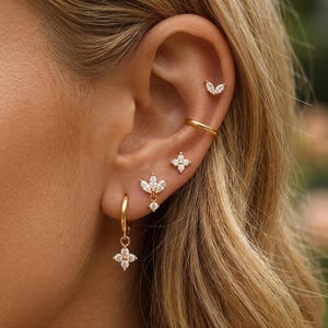 Op de afbeelding: Een set goudkleurige oorbellen en een goudkleurige oormanchet. De sieraden bestaan uit een kleine ring met een vierbladige bloemhanger, een driebladige bladstud met een bungelende steen, een vierpuntige sterstud, een effen gouden bandje.