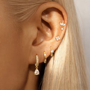 Könnte beinhalten: Nahaufnahme eines Ohrs, das mit mehreren goldenen Ohrringen geschmückt ist. Die Ohrringe umfassen kleine Creolen, einen tropfenförmigen Anhänger und Ohrstecker mit klaren Edelsteinen. Das Schmuckstück ist vor blondem Haar platziert.