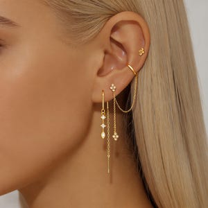 May include: A collection of gold-colored earrings and ear cuffs. The earrings feature various designs, including dangling chains, geometric shapes, and floral accents. The ear cuffs have a simple, elegant design. The jewelry is worn on a person's ear.
