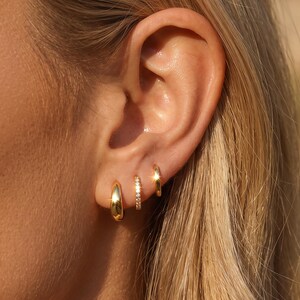 Peut inclure: Un ensemble de trois boucles d'oreilles créoles dorées portées sur un seul lobe d'oreille. La première est une créole lisse, épaisse et polie, la deuxième est une fine créole incrustée de petites pierres claires, et la troisième est une simple créole dorée.