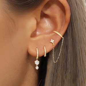 Può includere: Una collezione di orecchini dorati, tra cui orecchini a cerchio e un orecchino a doppia goccia con una pietra trasparente. Una catena collega un orecchino a forma di fiore a un ear cuff curvo. Gli orecchini sono incastonati con piccole pietre scintillanti.