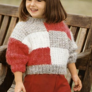 Op de afbeelding: Een patchwork trui in rood, wit en grijs. De trui heeft een ronde hals en lange mouwen. De mouwen en een deel van het lichaam zijn rood. De andere delen zijn wit en grijs. Het kind draagt de trui.